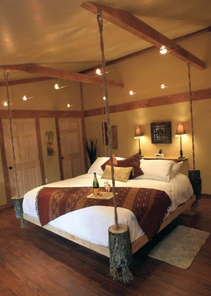 Rustic hanging bed with log-style supports, wooden beams, and warm lighting in a cozy bedroom.