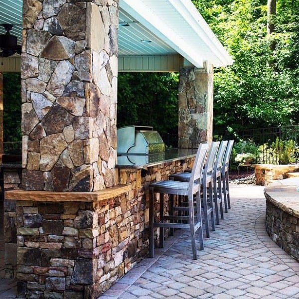 Covered outdoor stone bar with metal barstools and built-in grill on a cobblestone patio.