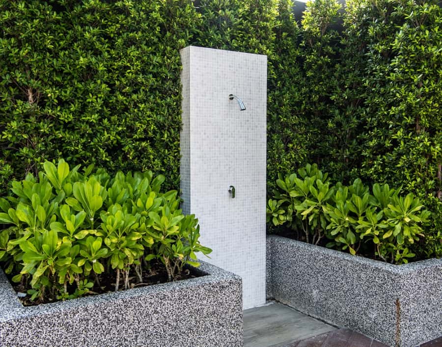 Minimalist outdoor shower with white mosaic tiles, surrounded by dense green hedges and planters.