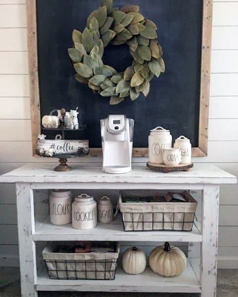 Rustic coffee bar with a white wooden table, shiplap walls, chalkboard, and decorative green wreath.