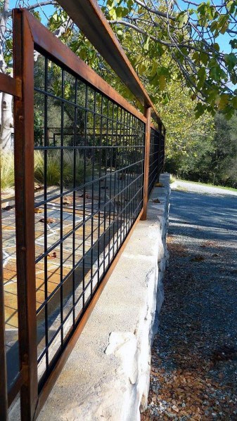 A metal fence crowns a stone wall amid trees and a sunlit pathway, creating a serene outdoor setting