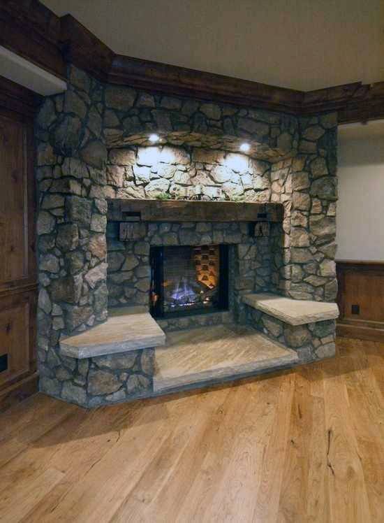 Rustic stone corner fireplace with built-in seating and wood mantel on hardwood flooring.