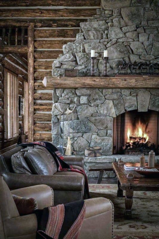 Rustic log cabin living room with large stone fireplace, wooden mantel, and cozy leather seating.