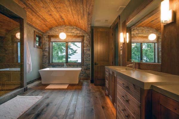 Rustic bathroom with wooden floors, stone walls, and a freestanding tub by expansive windows