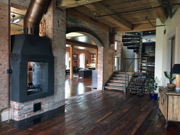 Rustic loft with exposed brick, wooden beams, spiral staircase, and double-sided fireplace.