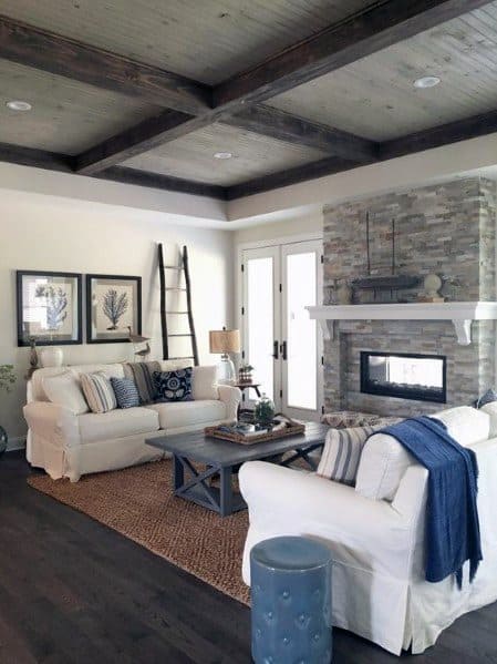 Cozy living room with rustic coffered wood ceiling, stone fireplace, white sofas, and blue accents.