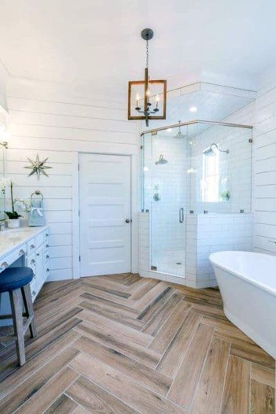 A modern corner bathroom with a glass shower, freestanding tub, wooden herringbone floor, and wooden pendant light