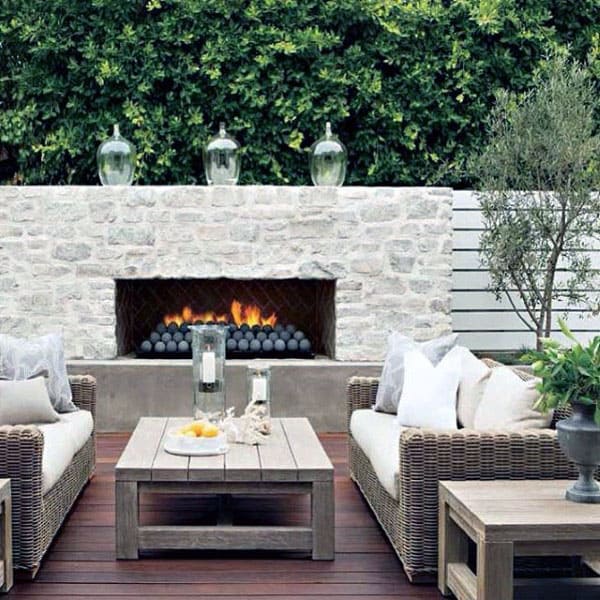 Cozy outdoor patio with wicker furniture, a lit stone fireplace, and greenery in the background