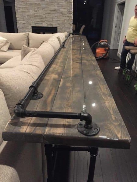Long wooden bar top with black industrial plumbing pipe supports, next to beige sectional sofa.
