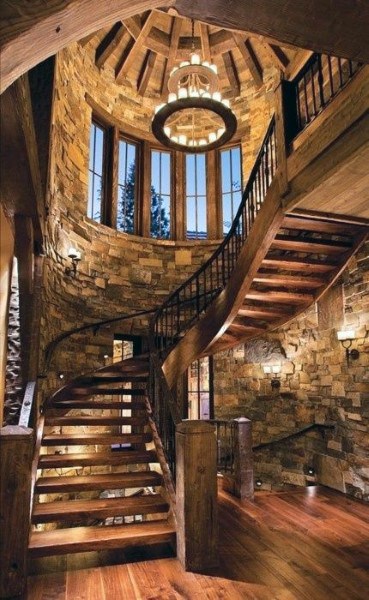 Rustic spiral staircase with stone walls, wooden steps, and large windows; a circular chandelier illuminates the staircase