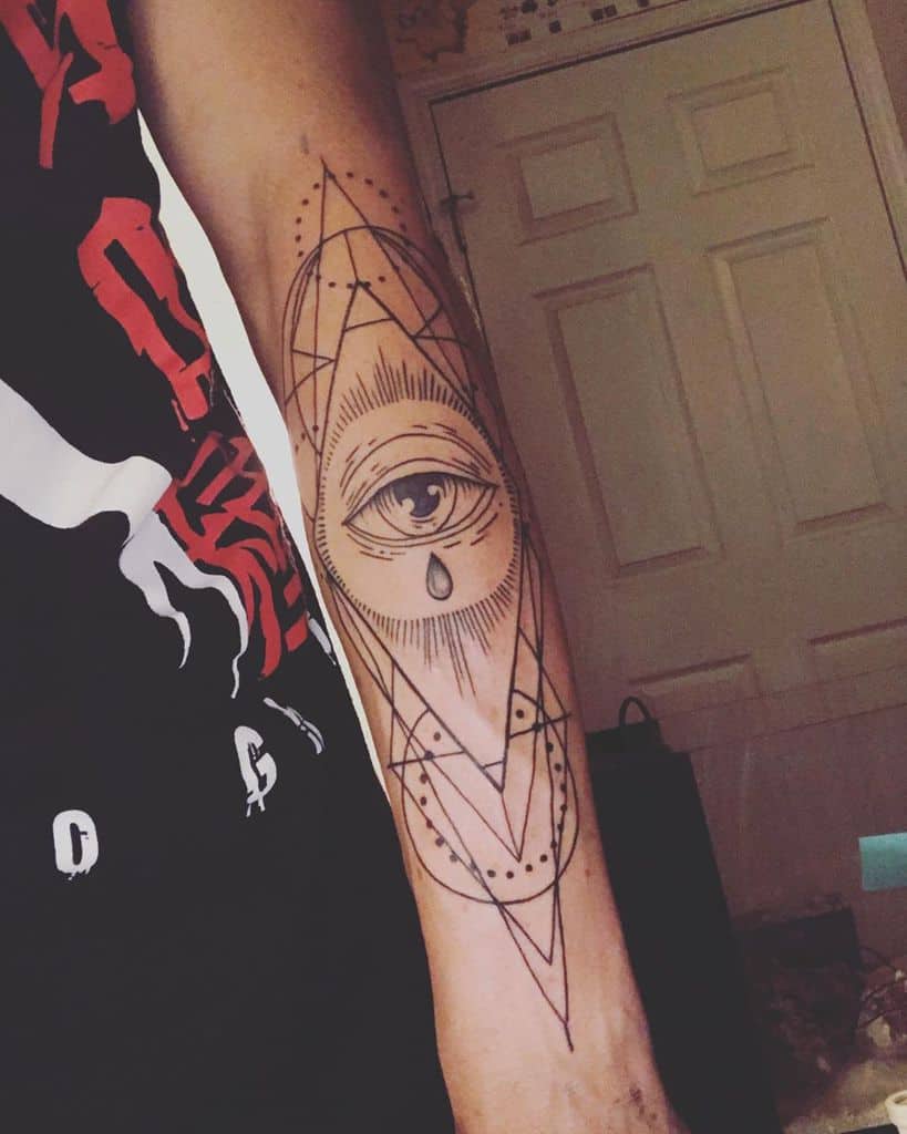 Tattoo of an all-seeing eye with a teardrop, surrounded by geometric shapes and fine line details, inked in black on the forearm.