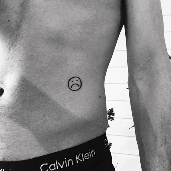 Minimalistic tattoo of a sad face emoji outlined on the lower ribcage of a person's torso