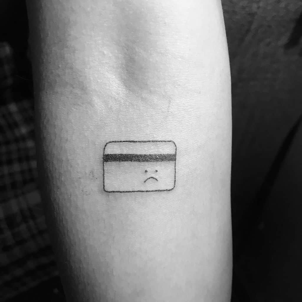 A minimalist black ink tattoo of a credit card with a sad face, symbolizing financial struggles in a humorous way.