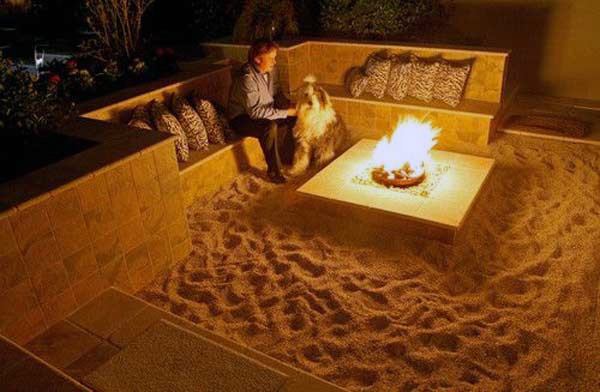 Person and dog relaxing by a glowing fire pit in a cozy outdoor seating area on sandy ground