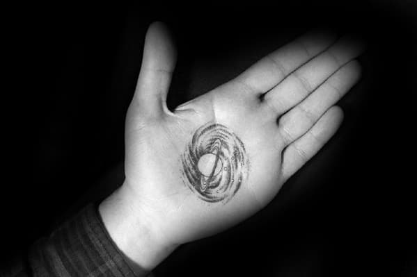 Sketch-style Saturn tattoo design for men inked on the palm, featuring swirling rings and rough cosmic lines in a bold black look.
