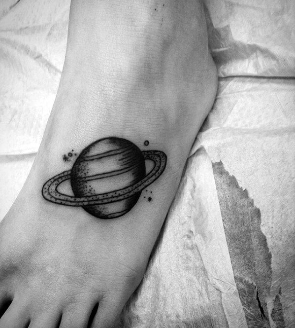 Small Saturn tattoo design for men placed on the foot, with bold black shading, ring details, and tiny surrounding stars.