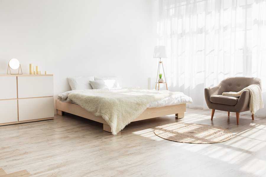 Bright, minimalist bedroom with a large bed, light wooden floor, a cozy armchair, and a small sideboard; sunlight streams through sheer curtains
