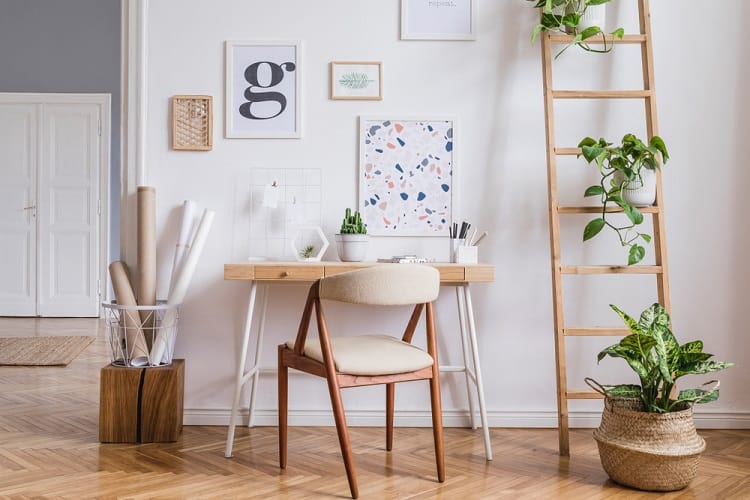 Minimalist workspace with a wooden desk, chair, ladder shelf, art prints, and potted plants on a herringbone floor