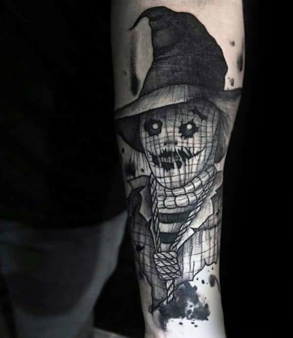 Black and grey scarecrow tattoo with button eyes, stitched smile, witch hat, rope neck, and ink splatter shading on the forearm.