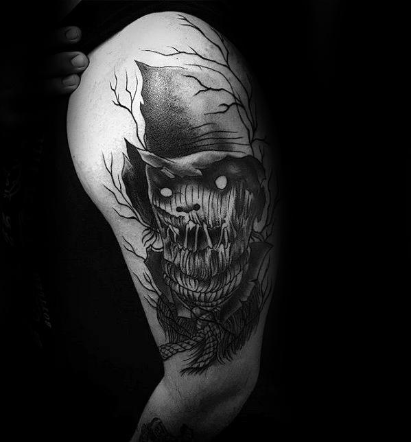 Creepy scarecrow tattoo on upper arm with glowing white eyes, stitched mouth, pointed hat, rope collar, and barren tree branches.