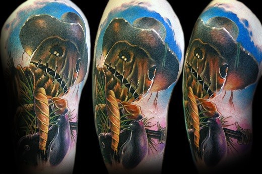 Vivid scarecrow tattoo with button eyes, stitched mouth, rope neck, and a wide-brim hat against a colorful twilight sky background.