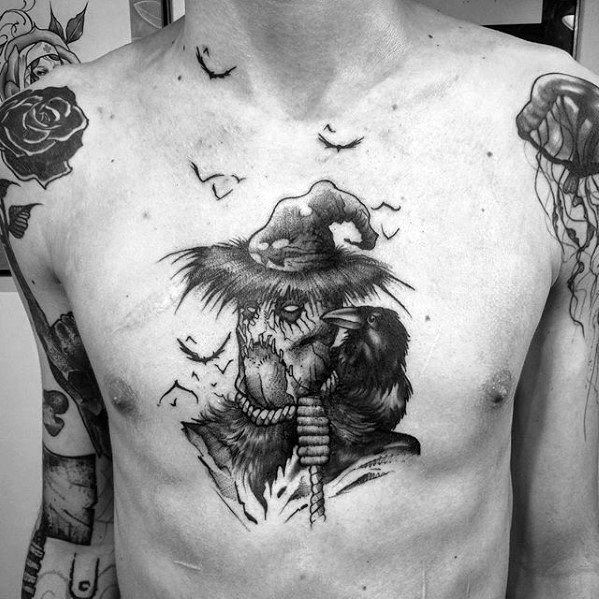 Black and grey chest tattoo of a stitched scarecrow with a witch hat, rope neck, perched crow, and flying birds in the background.
