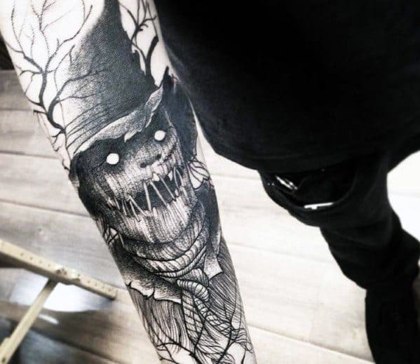 Forearm scarecrow tattoo with glowing white eyes, stitched grin, rope neck, wide-brim hat, and barren tree branches in blackwork style.