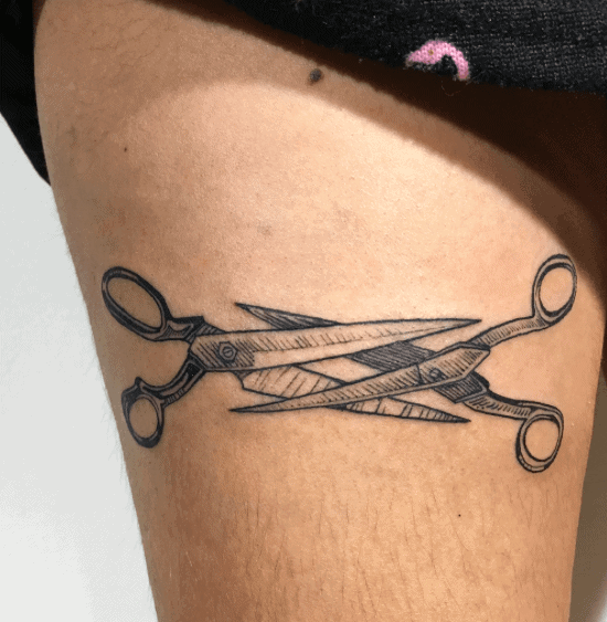 A black ink tattoo of two crossed scissors with detailed shading, creating a bold and sharp design on the upper thigh.