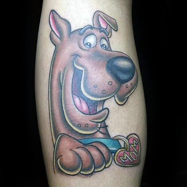 Tattoo of a happy Scooby-Doo holding a red heart-shaped tag that says “MOM,” with a cheerful and playful expression.