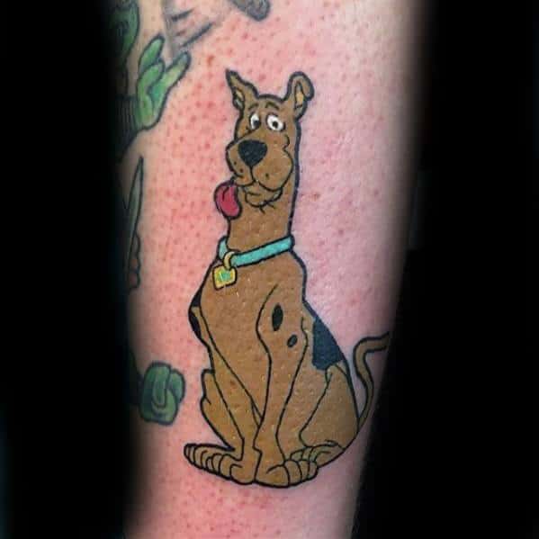 Tattoo of Scooby-Doo sitting with a goofy expression and tongue out, wearing his blue collar and gold diamond tag.