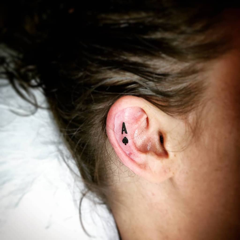 A small black tattoo of the Ace of Spades symbol, placed just behind the ear