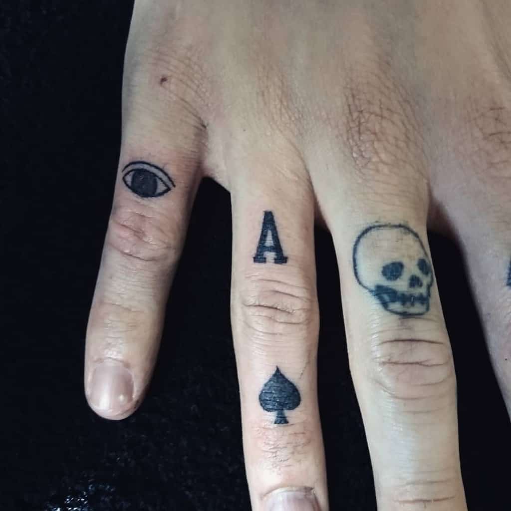 A tattoo design featuring an eye, the letter "A", and the Ace of Spades symbol on different fingers, with a skull tattoo placed on the pinky