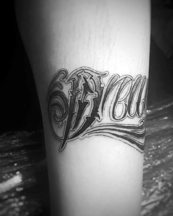 Forearm tattoo of the word “Dream” in bold gothic lettering, with intricate flourishes and stylized shading around the letters.