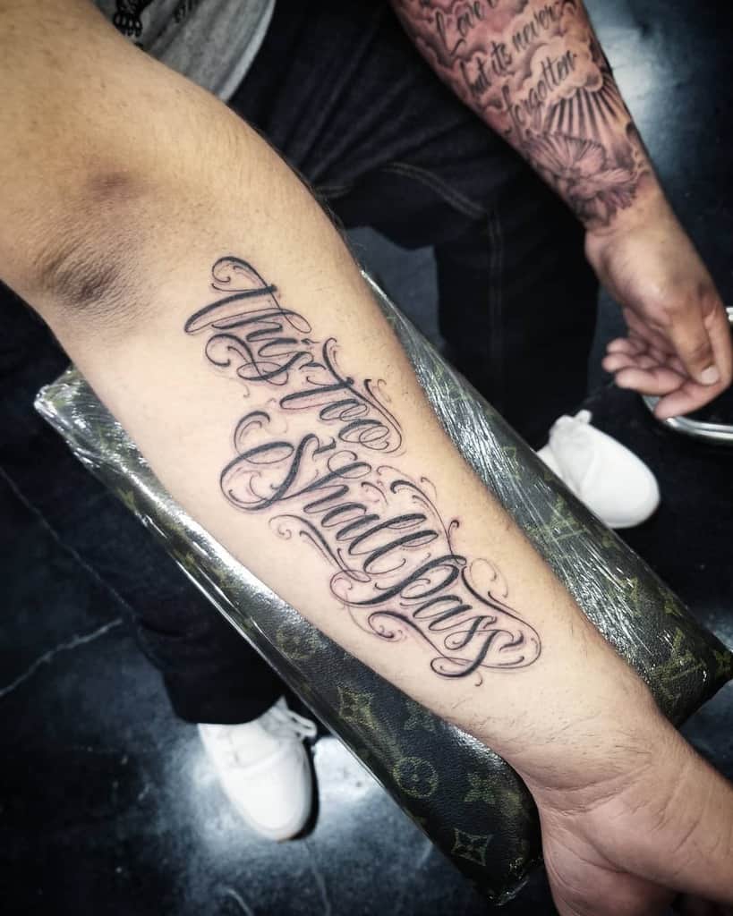 This "This Too Shall Pass" tattoo features an elegant script with decorative flourishes, giving it a refined and artistic look. The placement on the forearm makes it a bold and visible reminder of resilience and the transient nature of life.