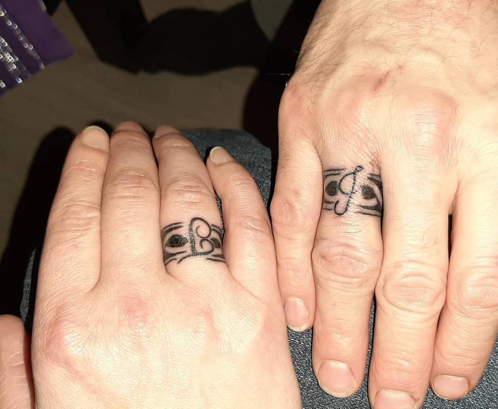 Two hands displaying matching wedding ring tattoos with artistic designs featuring letters and flowing lines