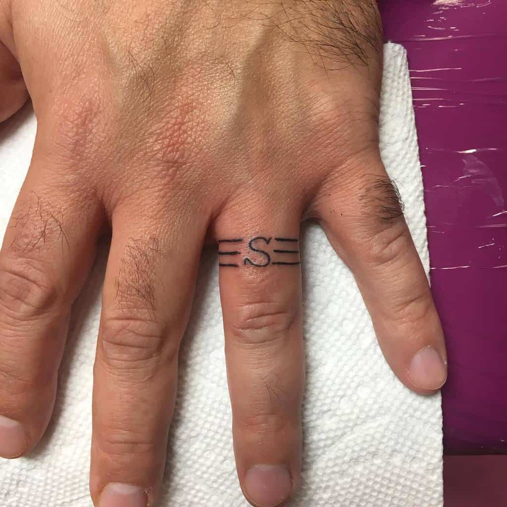 A close-up of a hand with a minimalistic wedding ring tattoo on the ring finger featuring an "S" letter with horizontal lines