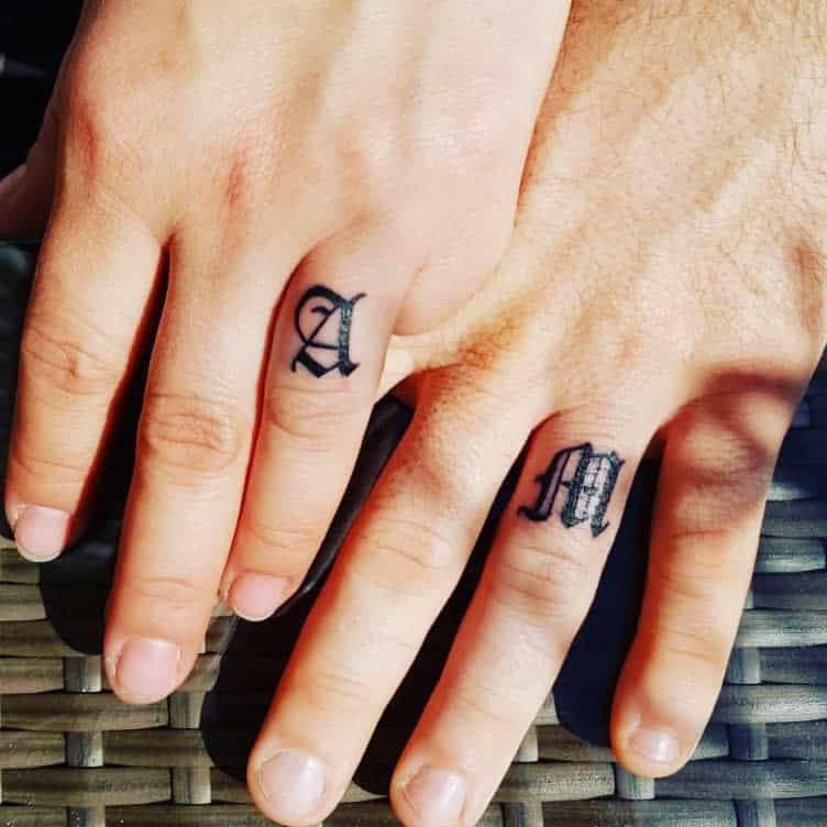 Two hands showing matching wedding ring tattoos with the letters "A" and "M" in an elegant, script font