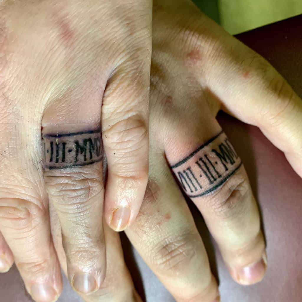Two hands with matching wedding ring tattoos featuring roman numeral designs, symbolizing a significant date or bond