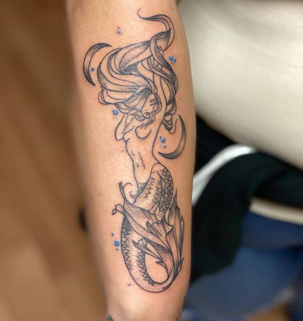 Elegant black and grey tattoo of a mermaid with flowing hair, detailed scales, and crescent moons, accented with small blue water droplets.