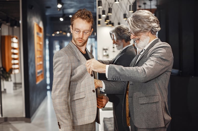 A grey-haired tailor assists a customer in adjusting his plaid suit jacket in a modern, well-lit store