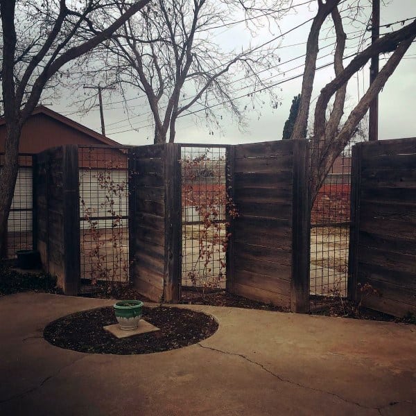 Rustic wooden fence with wire mesh panels enclosing a backyard patio area with trees.
