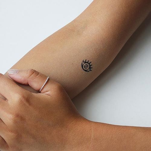 Small minimalist tattoo of an all-seeing eye with bold black lines and a curved design, inked on the inner forearm.