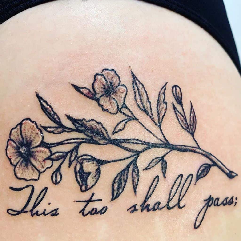 This tattoo features the phrase "This too shall pass" in elegant cursive, intertwined with a detailed floral design. The flowers and leaves add a delicate yet strong aesthetic, reinforcing the message of resilience and change.