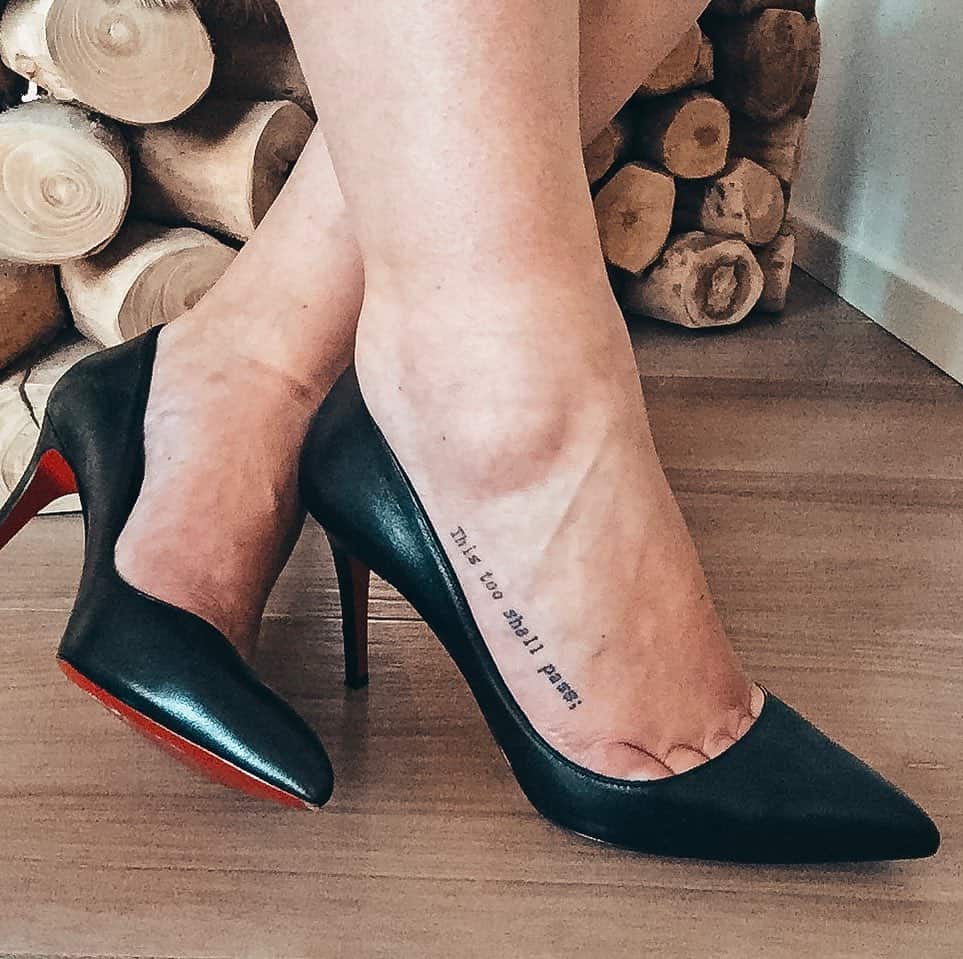 The image shows a tattoo of the phrase "This too shall pass" on the person's ankle, paired with high-heeled shoes.