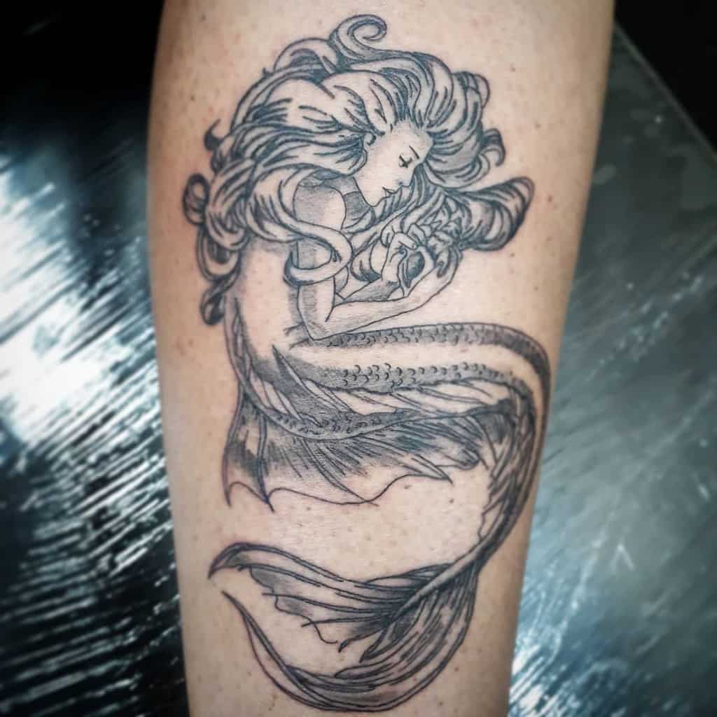 Black and grey tattoo of a mermaid with flowing hair, curled up while holding a seashell, with delicate shading and intricate details.