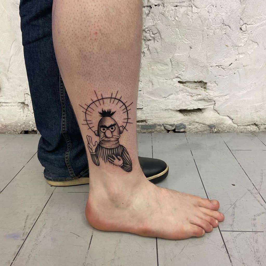 A black ink tattoo of a cartoon character with a serious expression, depicted as a saint with a halo and a blessing gesture, placed on the lower leg.