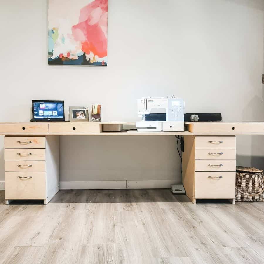 Modern sewing workstation featuring a minimalist wooden desk with multiple drawers for storage, a sewing machine setup, and a vibrant wall art centerpiece
