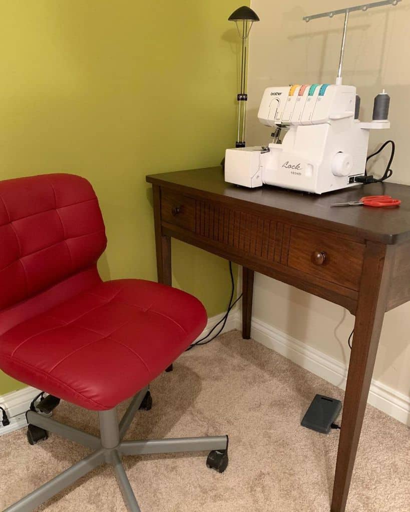 Compact sewing corner with a dark wood table featuring a Brother serger, paired with a bold red cushioned rolling chair, set against a vibrant green accent wall