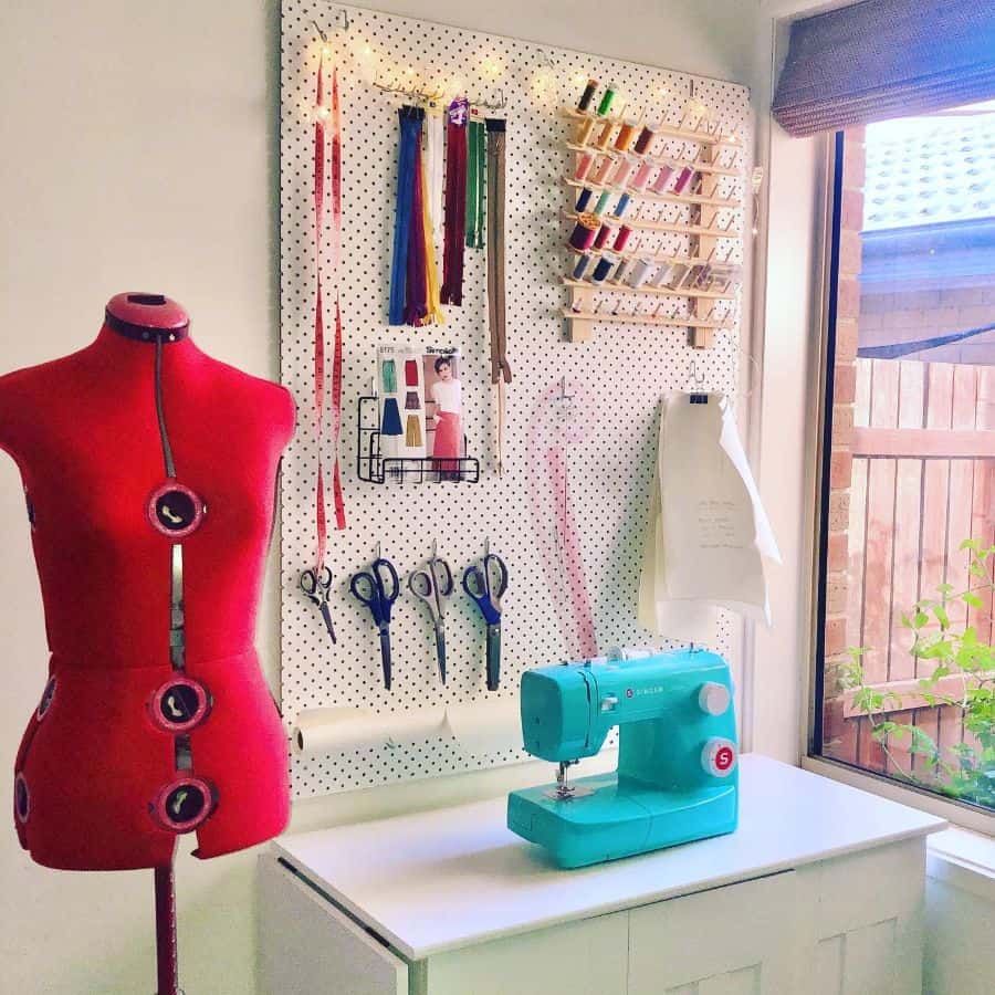 Colorful sewing area with a red dress form, teal sewing machine, organized pegboard, and a bright window view