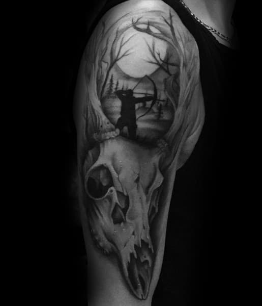 Black and grey upper arm tattoo of a deer skull with antlers forming a forest scene, featuring a bowhunter silhouette aiming under a full moon.